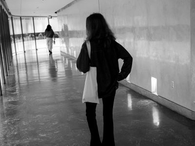 A person with a tote bag walks down a hallway, with another person visible farther ahead.