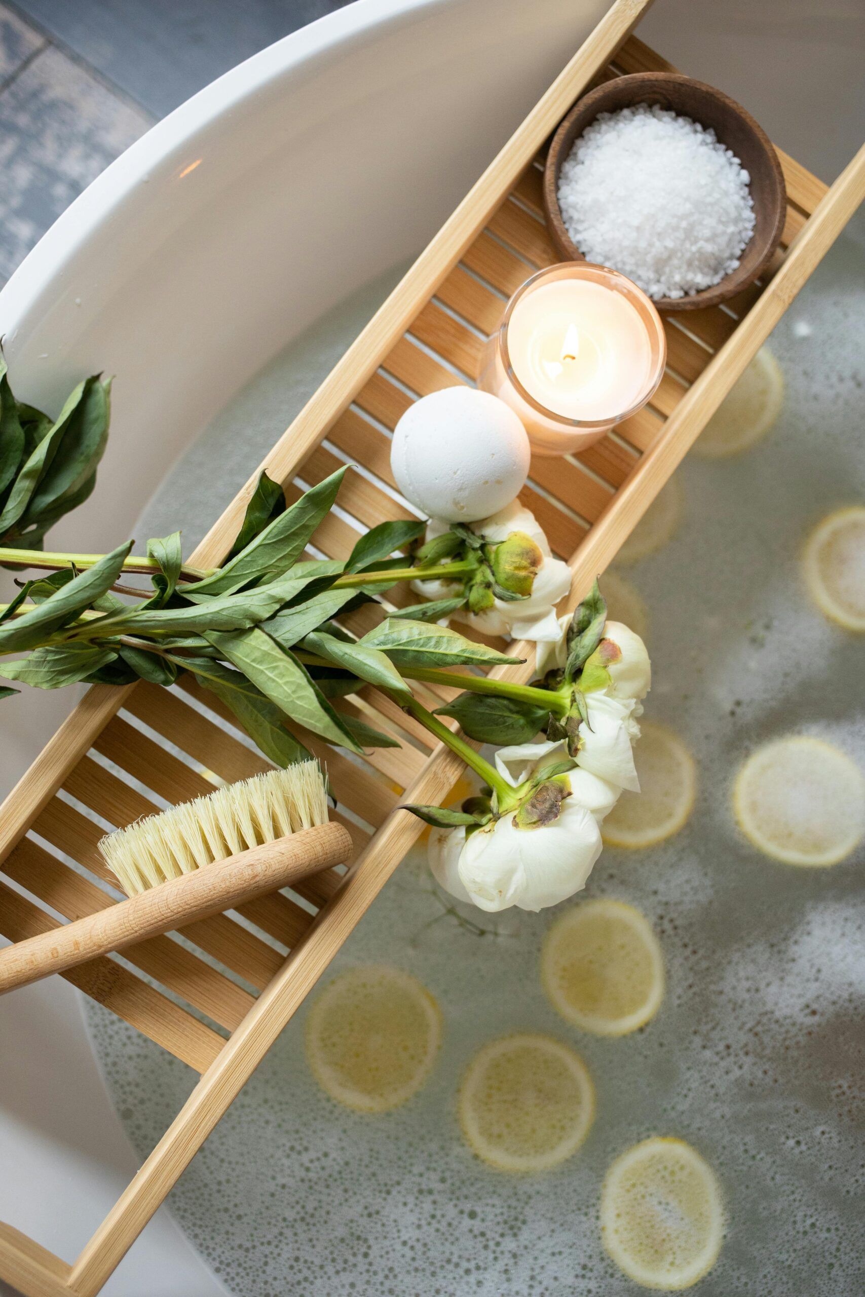 Bathtub with lemon slices, a tray holding peonies, a candle, bath salt, a brush, and a bath bomb.