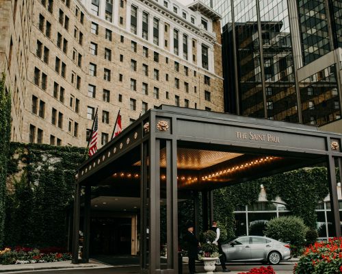 Landmark Tower Hotel entrance with "The Saint Paul" sign, flags, lit canopy, and greenery. A person stands near a parked car.