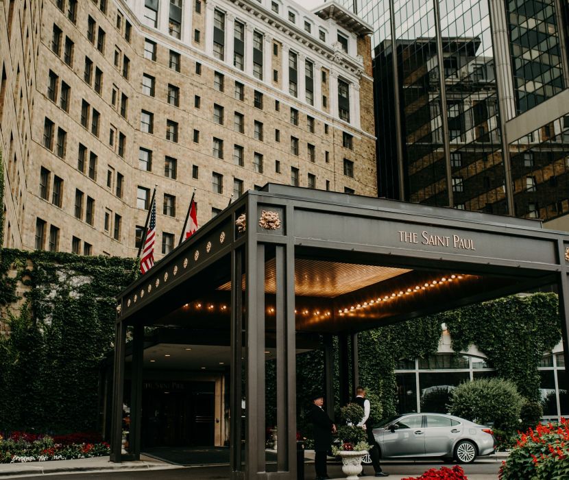 Landmark Tower Hotel entrance with "The Saint Paul" sign, flags, lit canopy, and greenery. A person stands near a parked car.