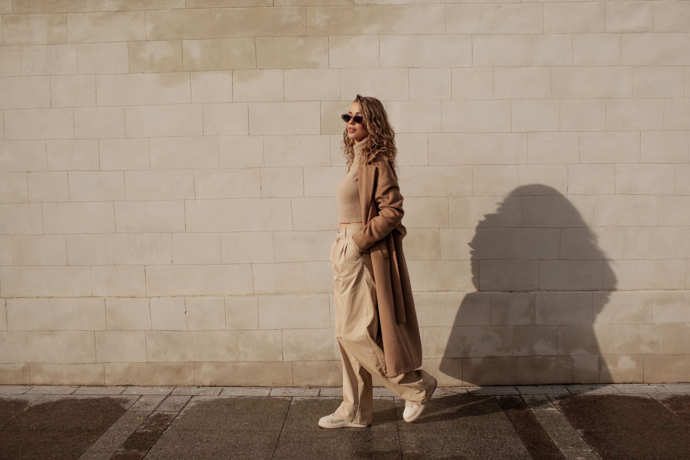 Landmark Tower Person in beige outfit and sunglasses walking along a sunlit brick wall.